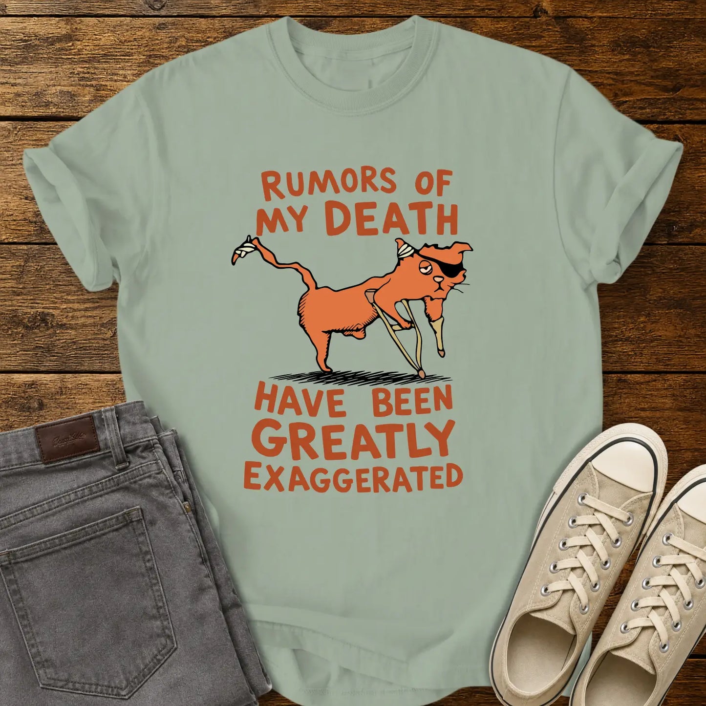 bay green T-shirt mockup featuring a humorous illustration of an orange cat with a peg leg, eye patch, and bandages alongside the phrase “RUMORS OF MY DEATH HAVE BEEN GREATLY EXAGGERATED” in retro orange lettering. The shirt is displayed on a rustic wood background alongside folded gray jeans and cream-colored canvas sneakers.