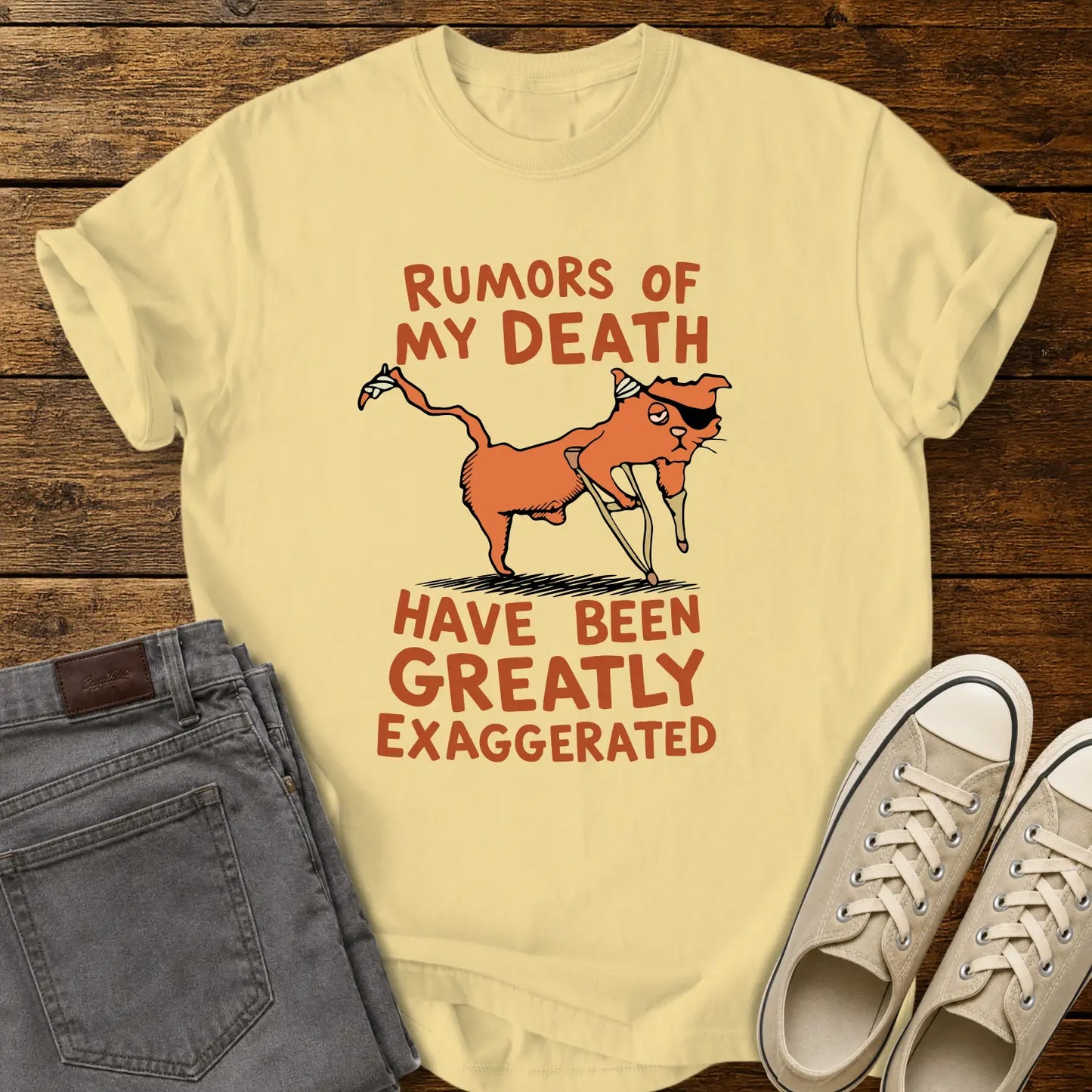 butter yellow T-shirt mockup featuring a humorous illustration of an orange cat with a peg leg, eye patch, and bandages alongside the phrase “RUMORS OF MY DEATH HAVE BEEN GREATLY EXAGGERATED” in retro orange lettering. The shirt is displayed on a rustic wood background alongside folded gray jeans and cream-colored canvas sneakers.