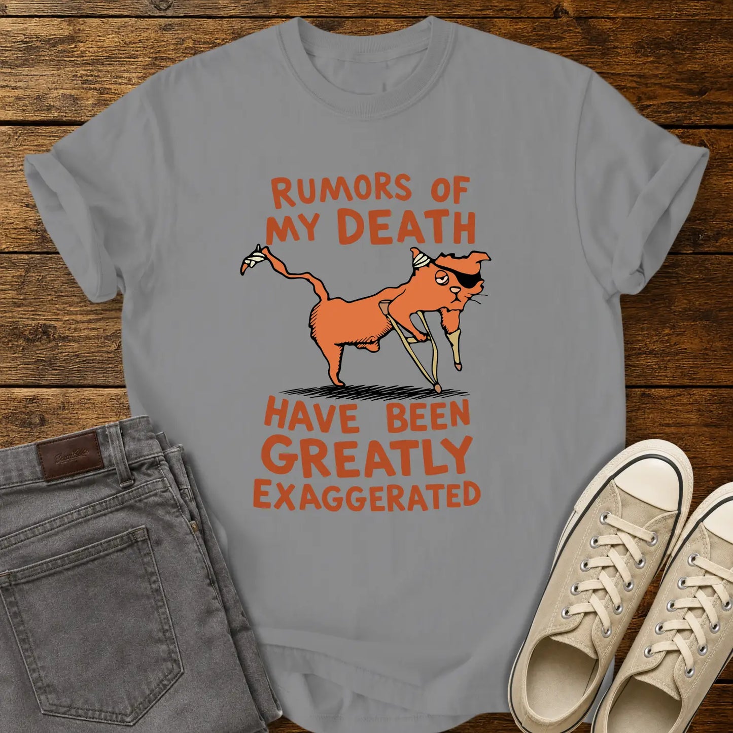 grey T-shirt mockup featuring a humorous illustration of an orange cat with a peg leg, eye patch, and bandages alongside the phrase “RUMORS OF MY DEATH HAVE BEEN GREATLY EXAGGERATED” in retro orange lettering. The shirt is displayed on a rustic wood background alongside folded gray jeans and cream-colored canvas sneakers.