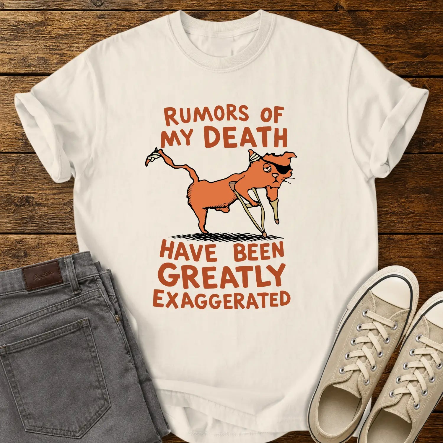 Ivory T-shirt mockup featuring a humorous illustration of an orange cat with a peg leg, eye patch, and bandages alongside the phrase “RUMORS OF MY DEATH HAVE BEEN GREATLY EXAGGERATED” in retro orange lettering. The shirt is displayed on a rustic wood background alongside folded gray jeans and cream-colored canvas sneakers.