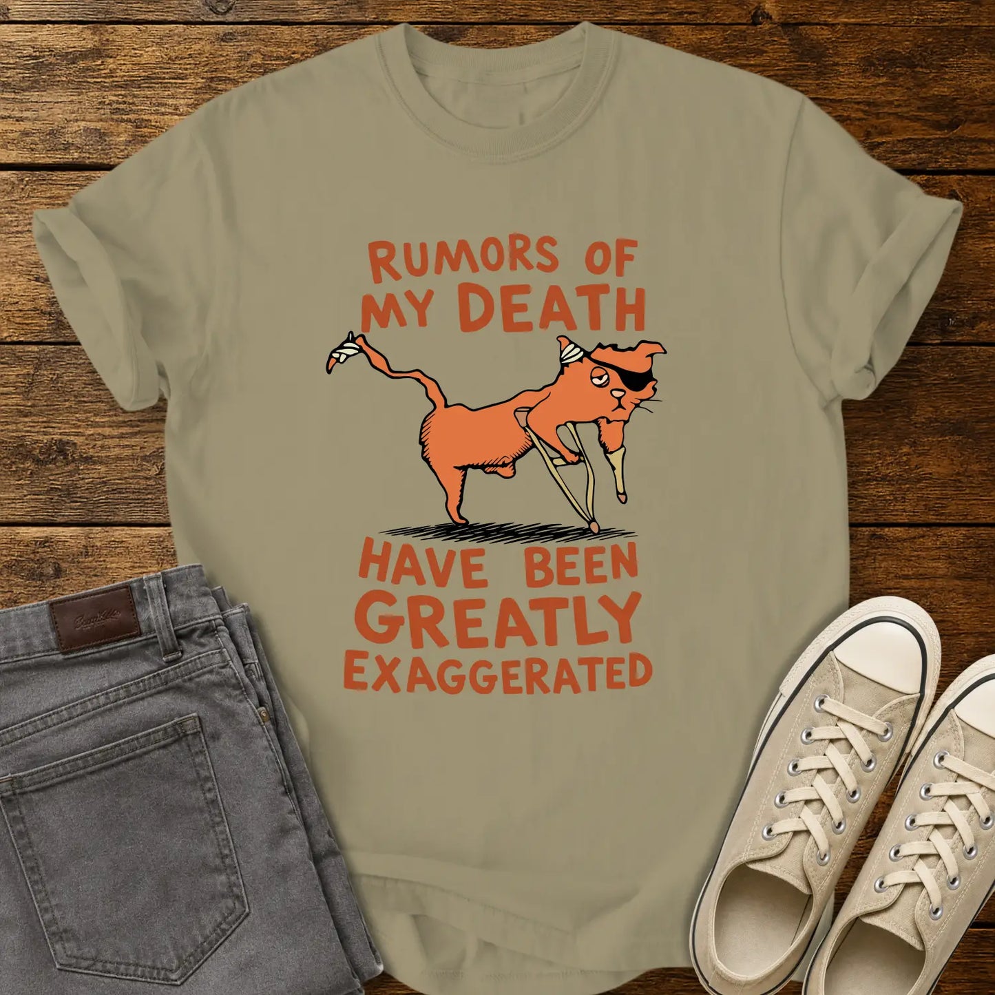 khaki T-shirt mockup featuring a humorous illustration of an orange cat with a peg leg, eye patch, and bandages alongside the phrase “RUMORS OF MY DEATH HAVE BEEN GREATLY EXAGGERATED” in retro orange lettering. The shirt is displayed on a rustic wood background alongside folded gray jeans and cream-colored canvas sneakers.