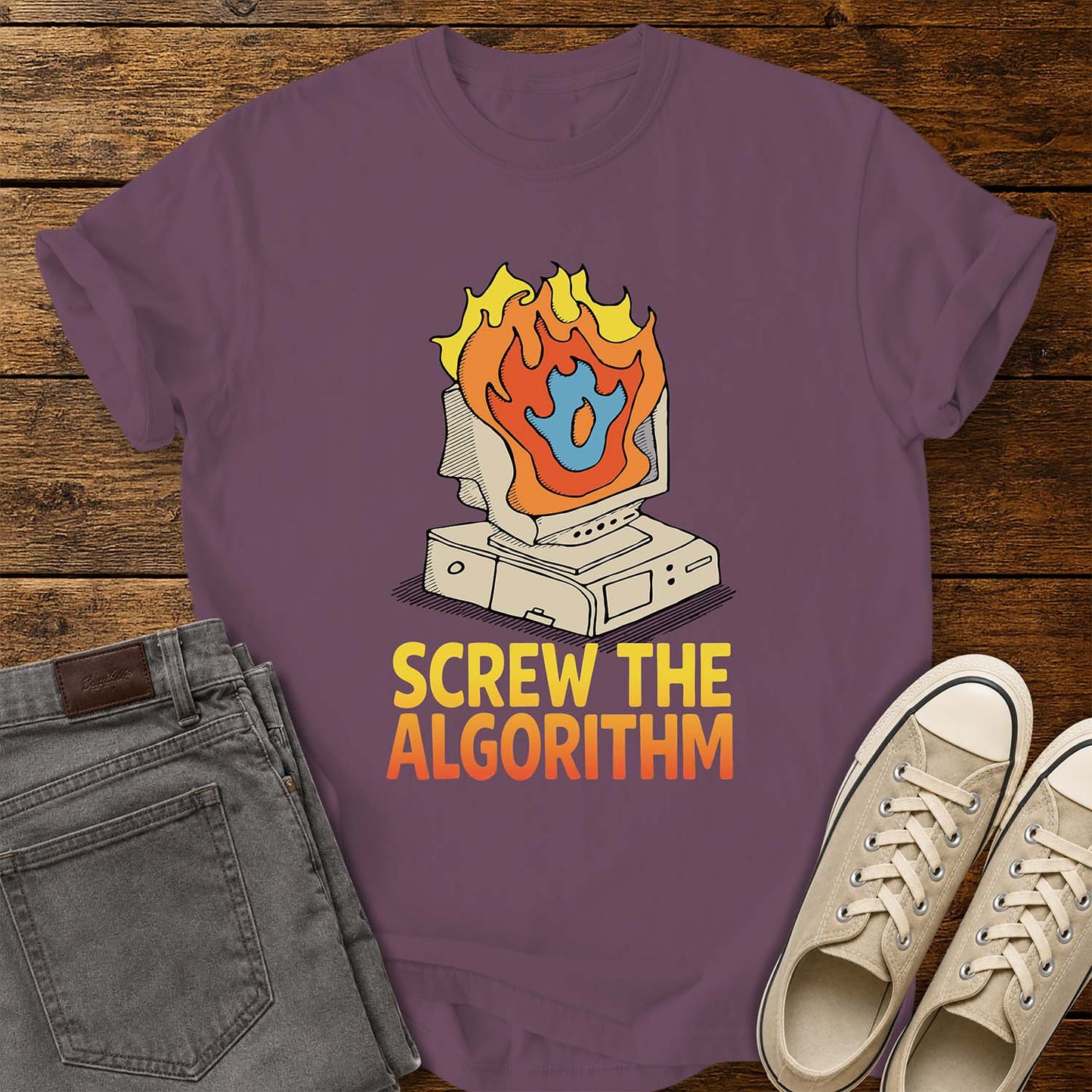 Berry purple T-shirt featuring a retro illustration of a vintage desktop computer with flames erupting from the monitor and a blue flame icon at the center. Beneath the graphic, bold yellow and orange text reads “SCREW THE ALGORITHM.” The shirt is displayed on a rustic wood plank background with folded gray jeans and a pair of cream canvas sneakers beside it.
