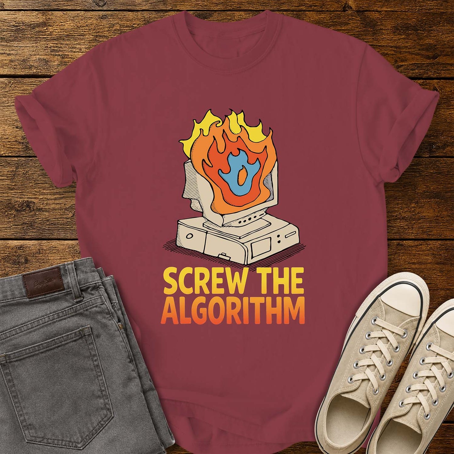 chili T-shirt featuring a retro illustration of a vintage desktop computer with flames erupting from the monitor and a blue flame icon at the center. Beneath the graphic, bold yellow and orange text reads “SCREW THE ALGORITHM.” The shirt is displayed on a rustic wood plank background with folded gray jeans and a pair of cream canvas sneakers beside it.
