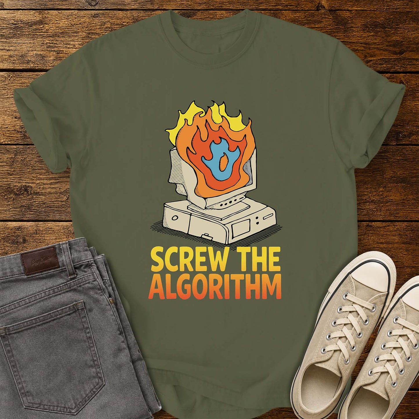 hemp green T-shirt featuring a retro illustration of a vintage desktop computer with flames erupting from the monitor and a blue flame icon at the center. Beneath the graphic, bold yellow and orange text reads “SCREW THE ALGORITHM.” The shirt is displayed on a rustic wood plank background with folded gray jeans and a pair of cream canvas sneakers beside it.