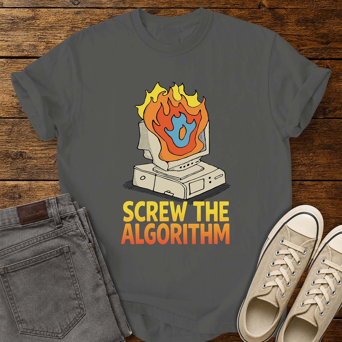 pepper T-shirt featuring a retro illustration of a vintage desktop computer with flames erupting from the monitor and a blue flame icon at the center. Beneath the graphic, bold yellow and orange text reads “SCREW THE ALGORITHM.” The shirt is displayed on a rustic wood plank background with folded gray jeans and a pair of cream canvas sneakers beside it.
