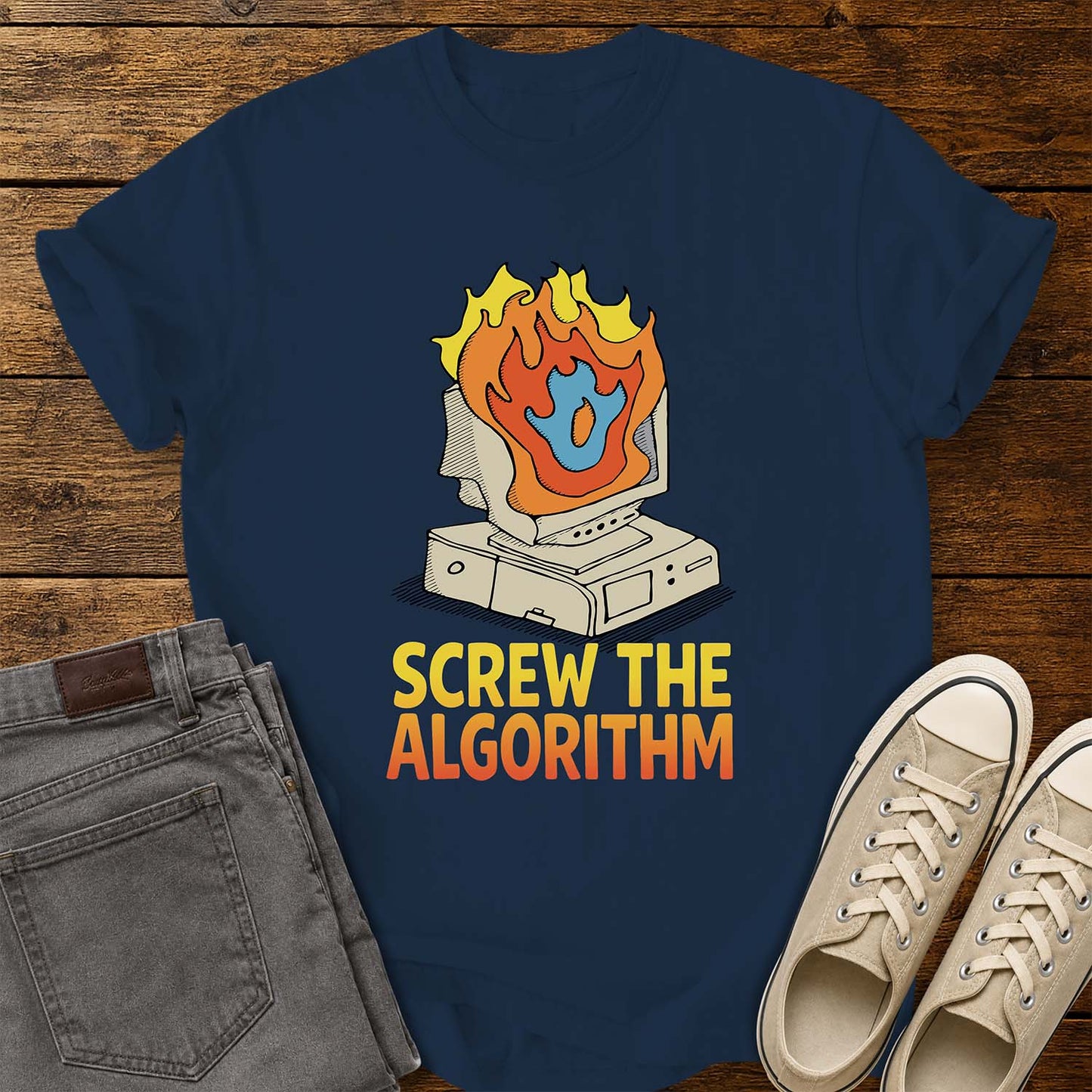 true navy T-shirt featuring a retro illustration of a vintage desktop computer with flames erupting from the monitor and a blue flame icon at the center. Beneath the graphic, bold yellow and orange text reads “SCREW THE ALGORITHM.” The shirt is displayed on a rustic wood plank background with folded gray jeans and a pair of cream canvas sneakers beside it.