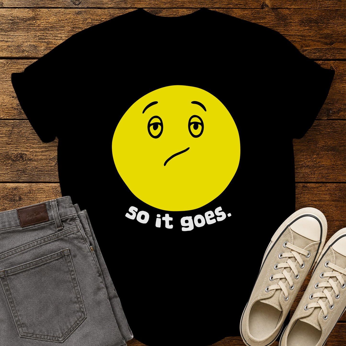 black flat-lay t-shirt mockup featuring a large yellow retro-style smiley face with a subtly uncertain expression above the phrase, "so it goes." in rounded white lowercase lettering. The minimalist vintage-inspired design is centered on the shirt, which is displayed on a rustic wood plank background alongside folded gray jeans and a pair of cream canvas sneakers.