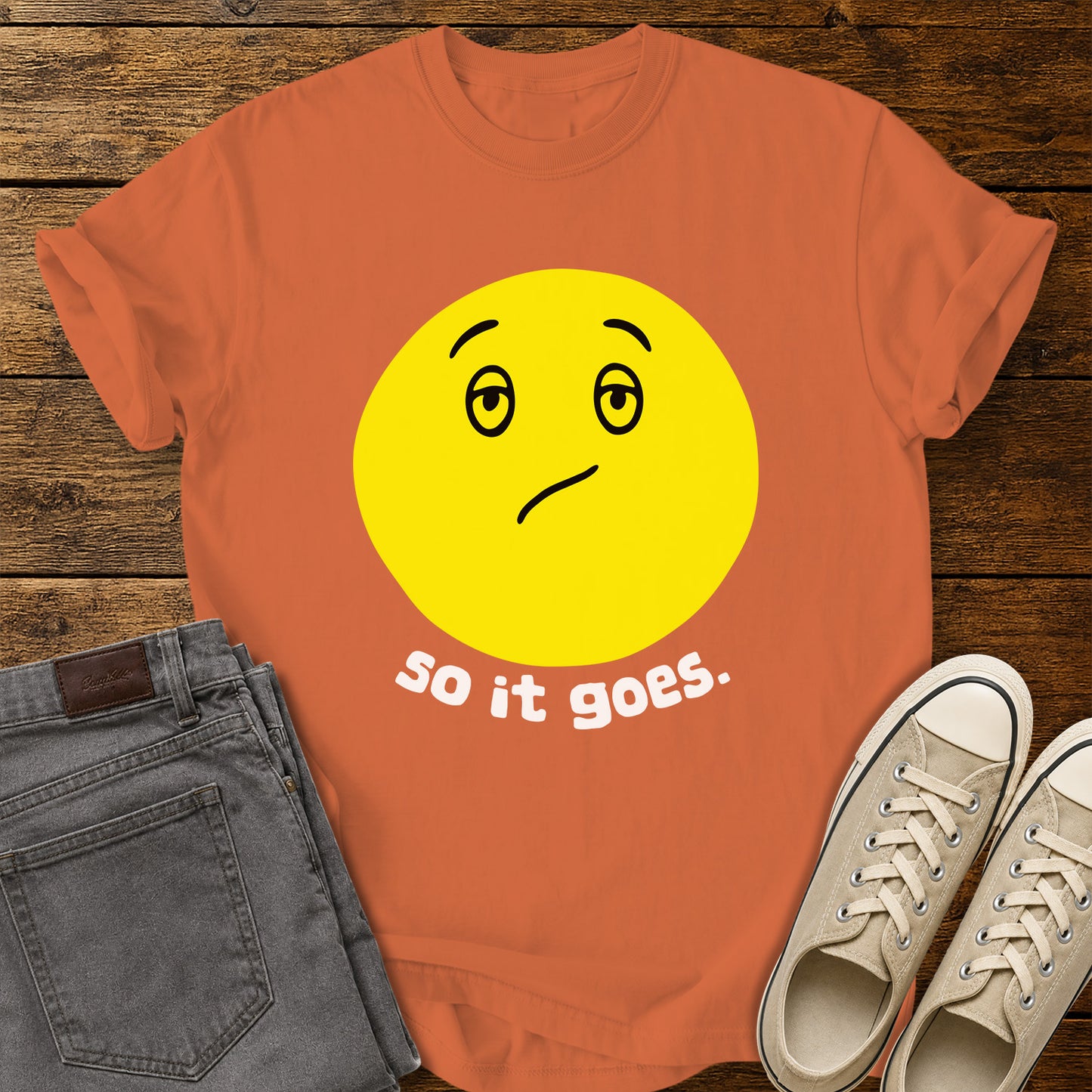 burnt orange flat-lay t-shirt mockup featuring a large yellow retro-style smiley face with a subtly uncertain expression above the phrase, "so it goes." in rounded white lowercase lettering. The minimalist vintage-inspired design is centered on the shirt, which is displayed on a rustic wood plank background alongside folded gray jeans and a pair of cream canvas sneakers.