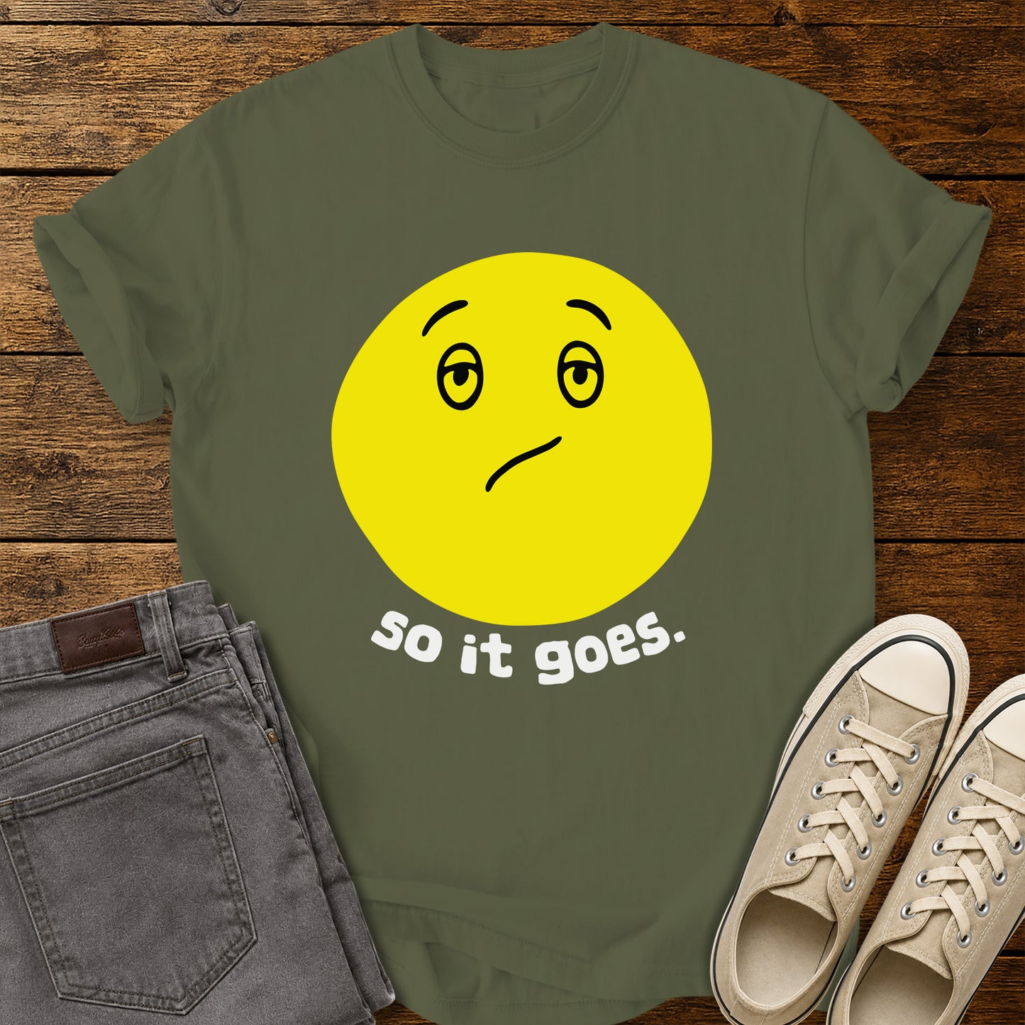 hemp green flat-lay t-shirt mockup featuring a large yellow retro-style smiley face with a subtly uncertain expression above the phrase, "so it goes." in rounded white lowercase lettering. The minimalist vintage-inspired design is centered on the shirt, which is displayed on a rustic wood plank background alongside folded gray jeans and a pair of cream canvas sneakers.