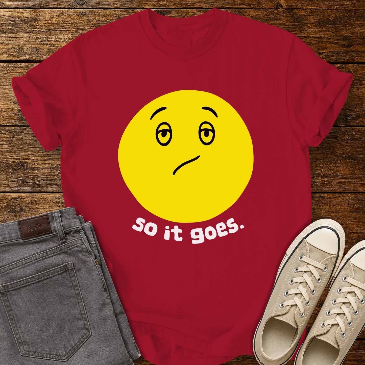 red flat-lay t-shirt mockup featuring a large yellow retro-style smiley face with a subtly uncertain expression above the phrase, "so it goes." in rounded white lowercase lettering. The minimalist vintage-inspired design is centered on the shirt, which is displayed on a rustic wood plank background alongside folded gray jeans and a pair of cream canvas sneakers.