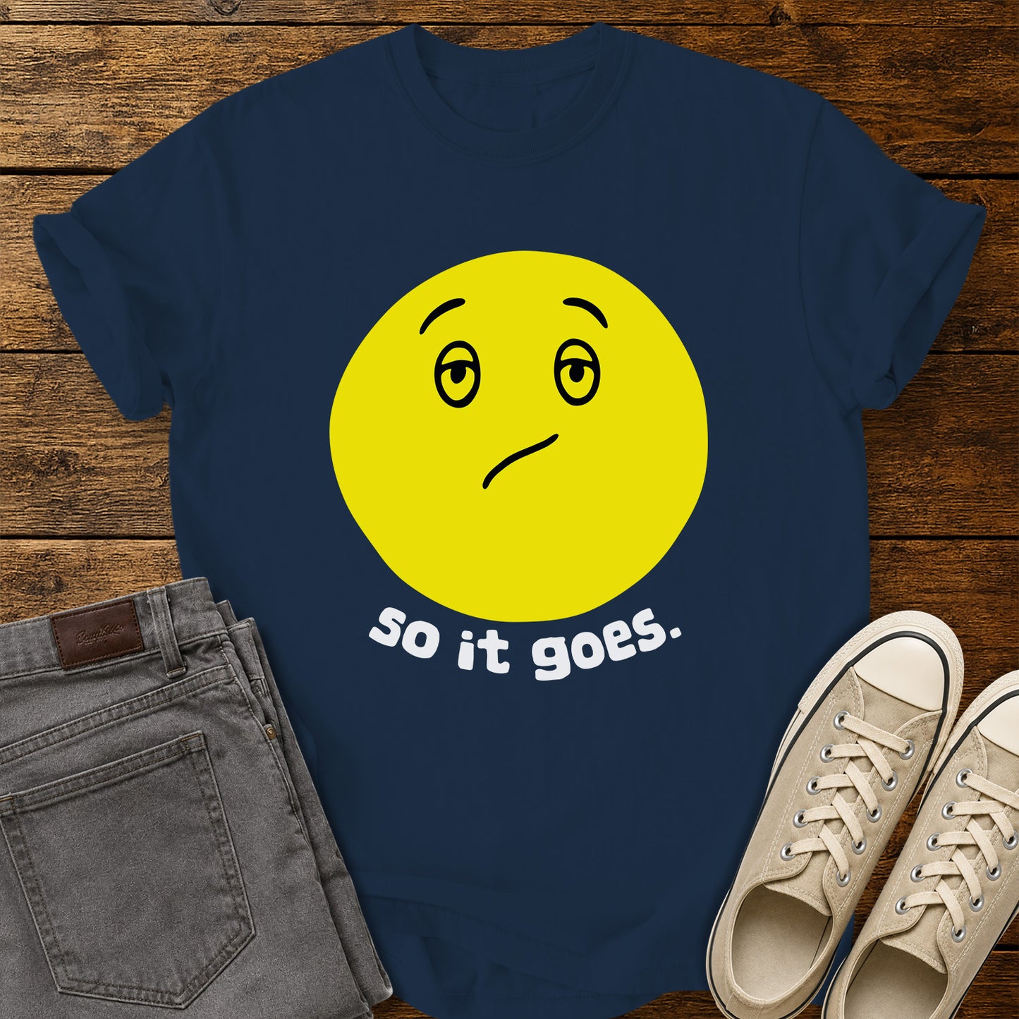 True navy flat-lay t-shirt mockup featuring a large yellow retro-style smiley face with a subtly uncertain expression above the phrase, "so it goes." in rounded white lowercase lettering. The minimalist vintage-inspired design is centered on the shirt, which is displayed on a rustic wood plank background alongside folded gray jeans and a pair of cream canvas sneakers.