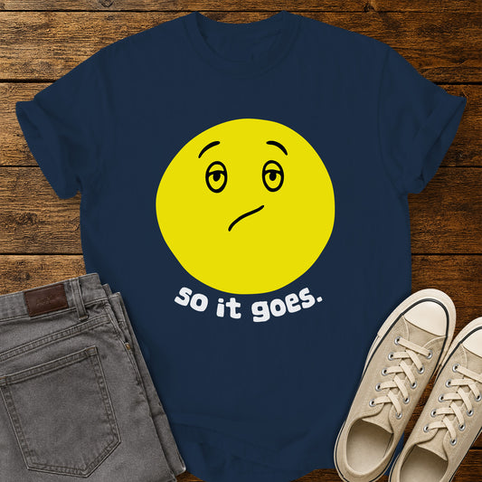 True navy flat-lay t-shirt mockup featuring a large yellow retro-style smiley face with a subtly uncertain expression above the phrase, "so it goes." in rounded white lowercase lettering. The minimalist vintage-inspired design is centered on the shirt, which is displayed on a rustic wood plank background alongside folded gray jeans and a pair of cream canvas sneakers.