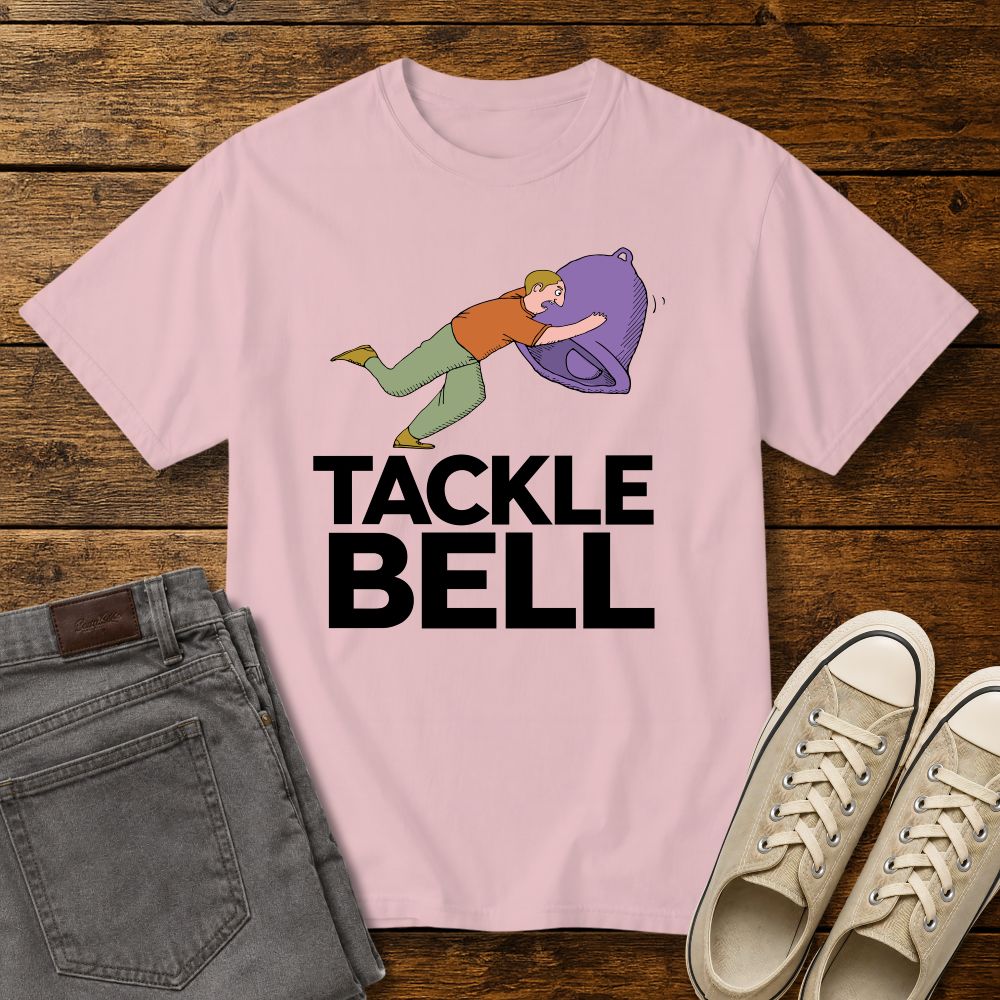 blossom pink color graphic T-shirt mockup featuring a cartoon-style man tackling a large bell with the phrase “Tackle Bell” in bold black lettering, styled with gray jeans and beige sneakers on a rustic wood background.