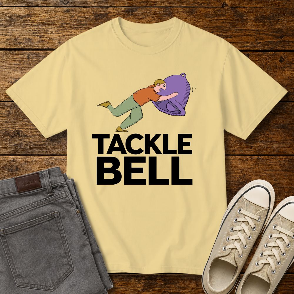 butter color graphic T-shirt mockup featuring a cartoon-style man tackling a large bell with the phrase “Tackle Bell” in bold black lettering, styled with gray jeans and beige sneakers on a rustic wood background.