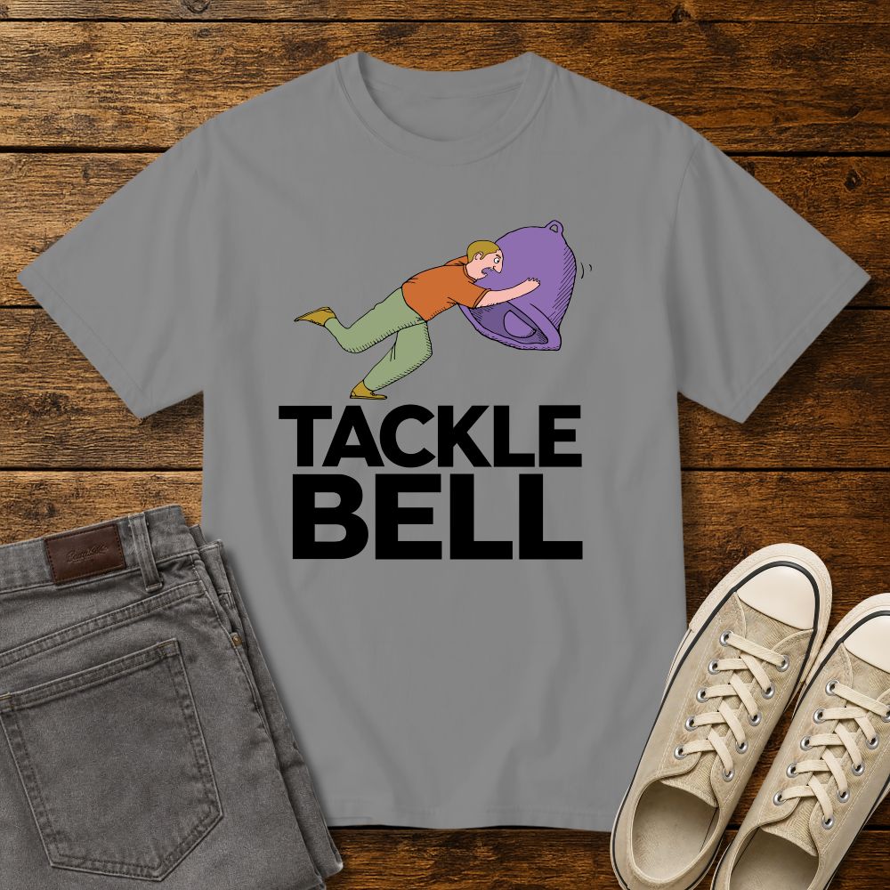 grey graphic T-shirt mockup featuring a cartoon-style man tackling a large bell with the phrase “Tackle Bell” in bold black lettering, styled with gray jeans and beige sneakers on a rustic wood background.
