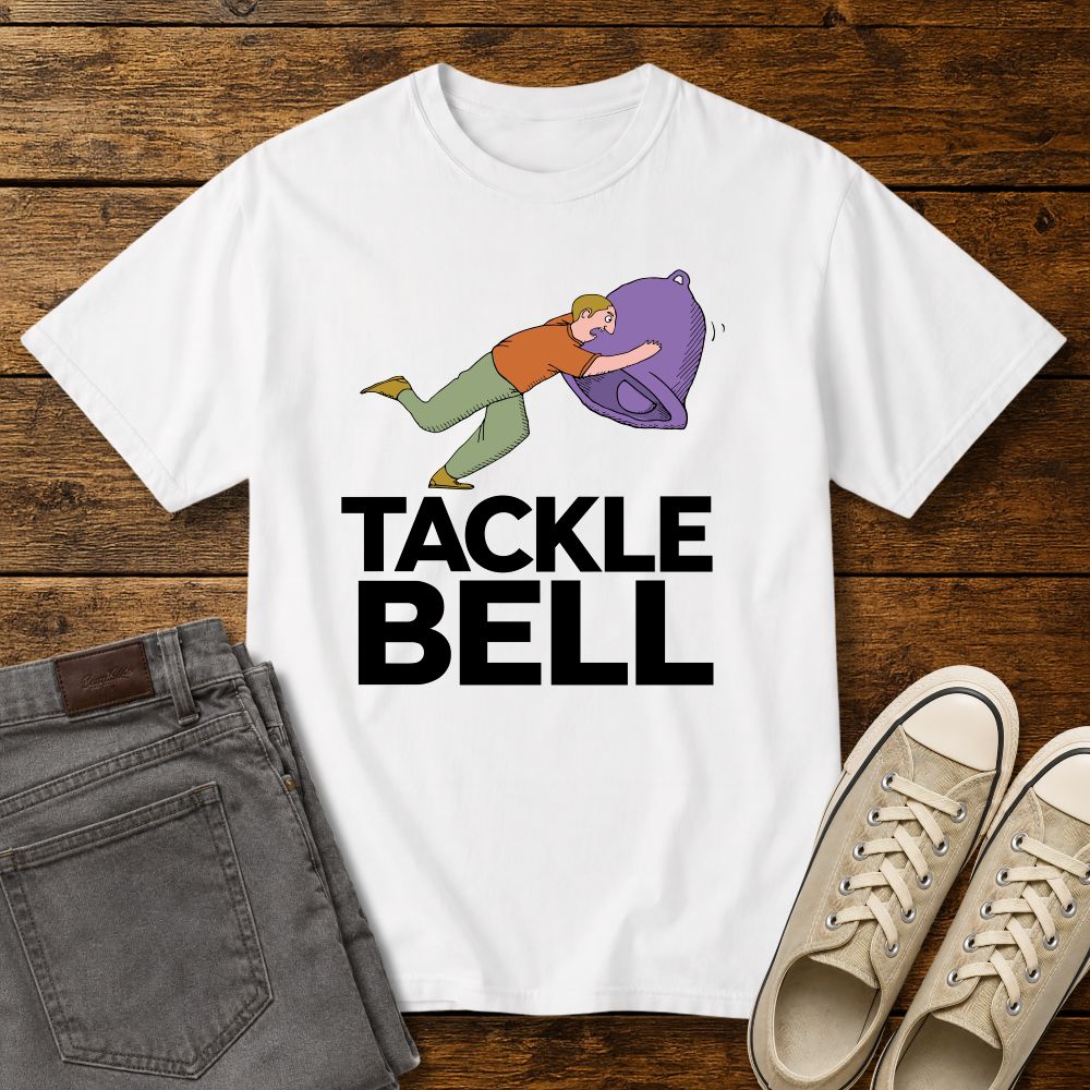 white  graphic T-shirt mockup featuring a cartoon-style man tackling a large bell with the phrase “Tackle Bell” in bold black lettering, styled with gray jeans and beige sneakers on a rustic wood background.