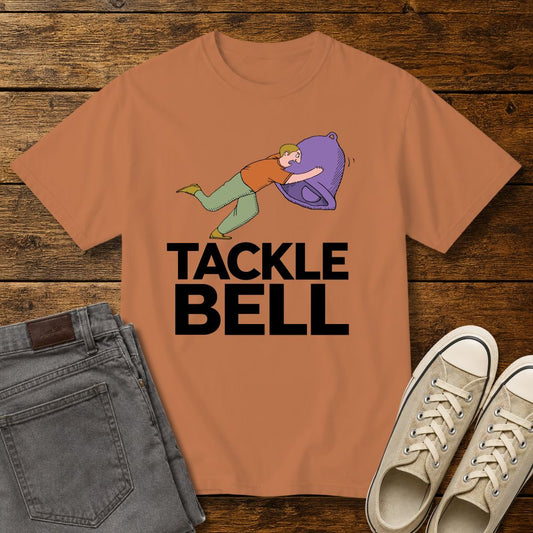 Yam-colored graphic T-shirt mockup featuring a cartoon-style man tackling a large bell with the phrase “Tackle Bell” in bold black lettering, styled with gray jeans and beige sneakers on a rustic wood background.