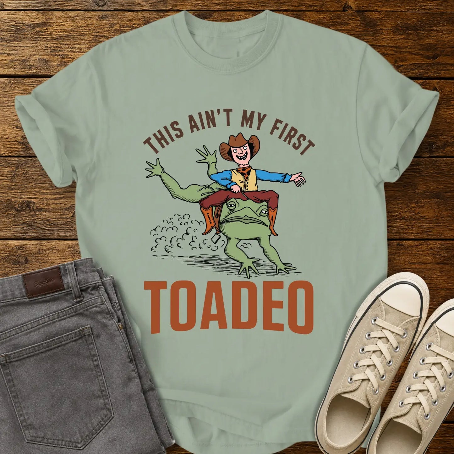 bay green T-shirt mockup featuring a humorous western design that reads “THIS AIN’T MY FIRST TOADEO.” The artwork shows a smiling cowboy riding a giant frog in a rodeo scene, with dust clouds behind them. The shirt is displayed on a rustic wood plank background alongside gray jeans and cream canvas sneakers.