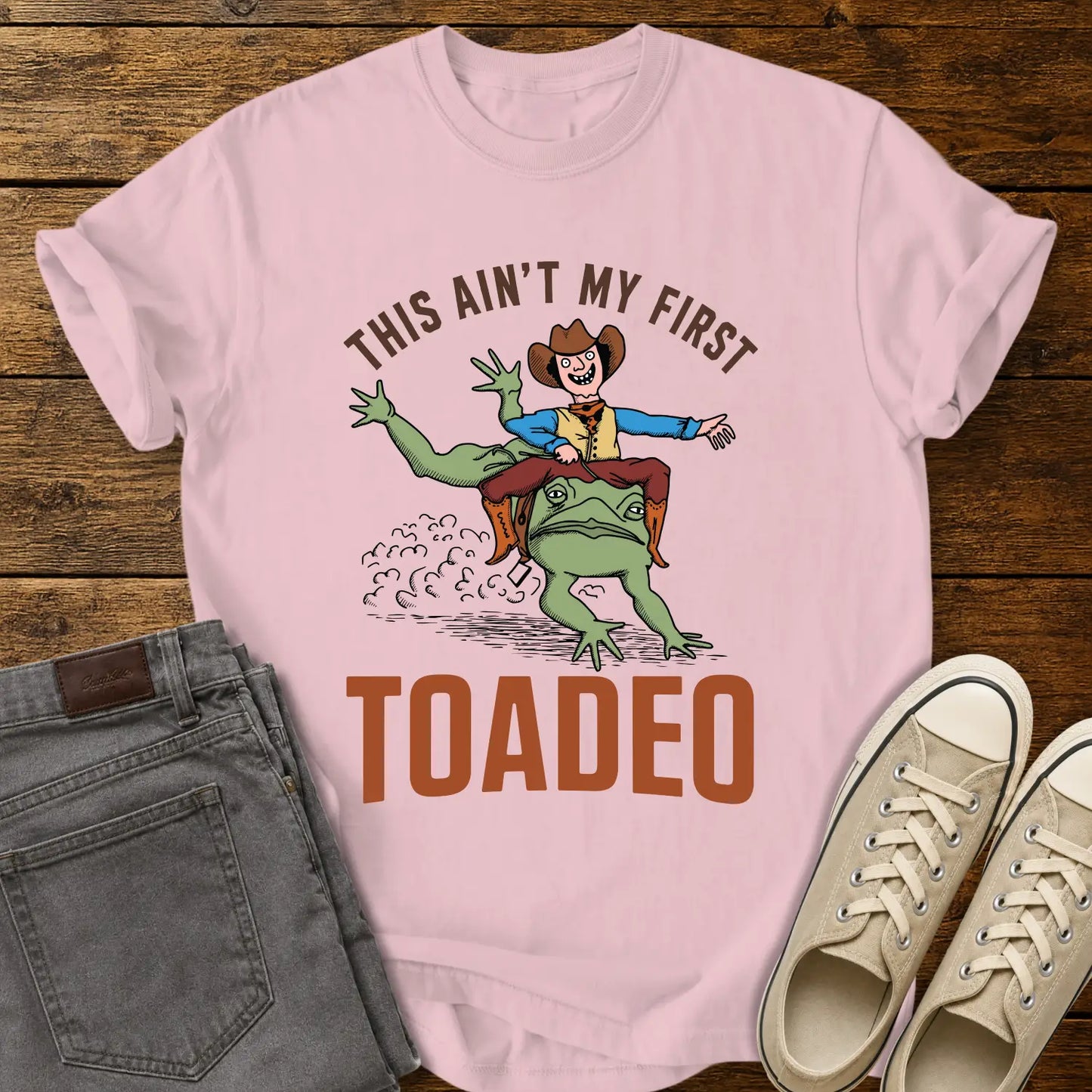 blossom pink T-shirt mockup featuring a humorous western design that reads “THIS AIN’T MY FIRST TOADEO.” The artwork shows a smiling cowboy riding a giant frog in a rodeo scene, with dust clouds behind them. The shirt is displayed on a rustic wood plank background alongside gray jeans and cream canvas sneakers.