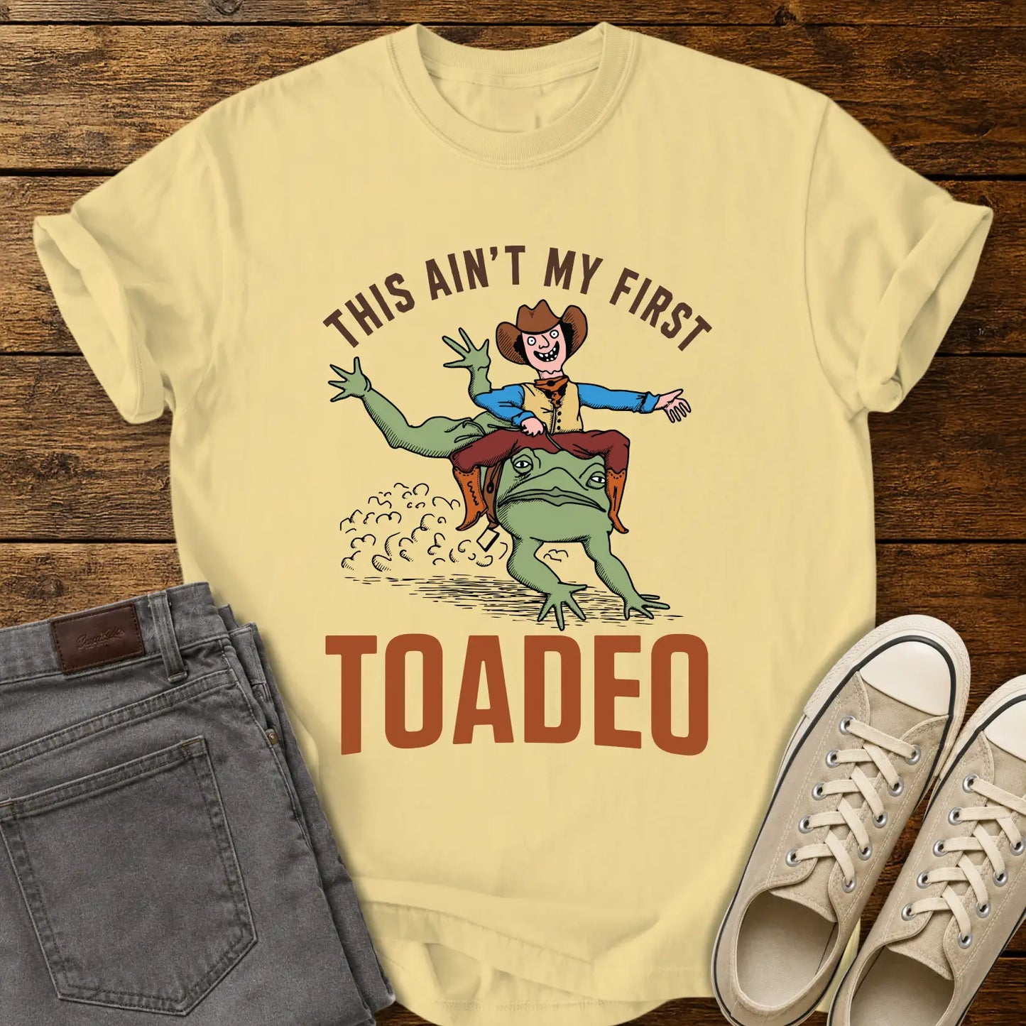 butter yellow T-shirt mockup featuring a humorous western design that reads “THIS AIN’T MY FIRST TOADEO.” The artwork shows a smiling cowboy riding a giant frog in a rodeo scene, with dust clouds behind them. The shirt is displayed on a rustic wood plank background alongside gray jeans and cream canvas sneakers.