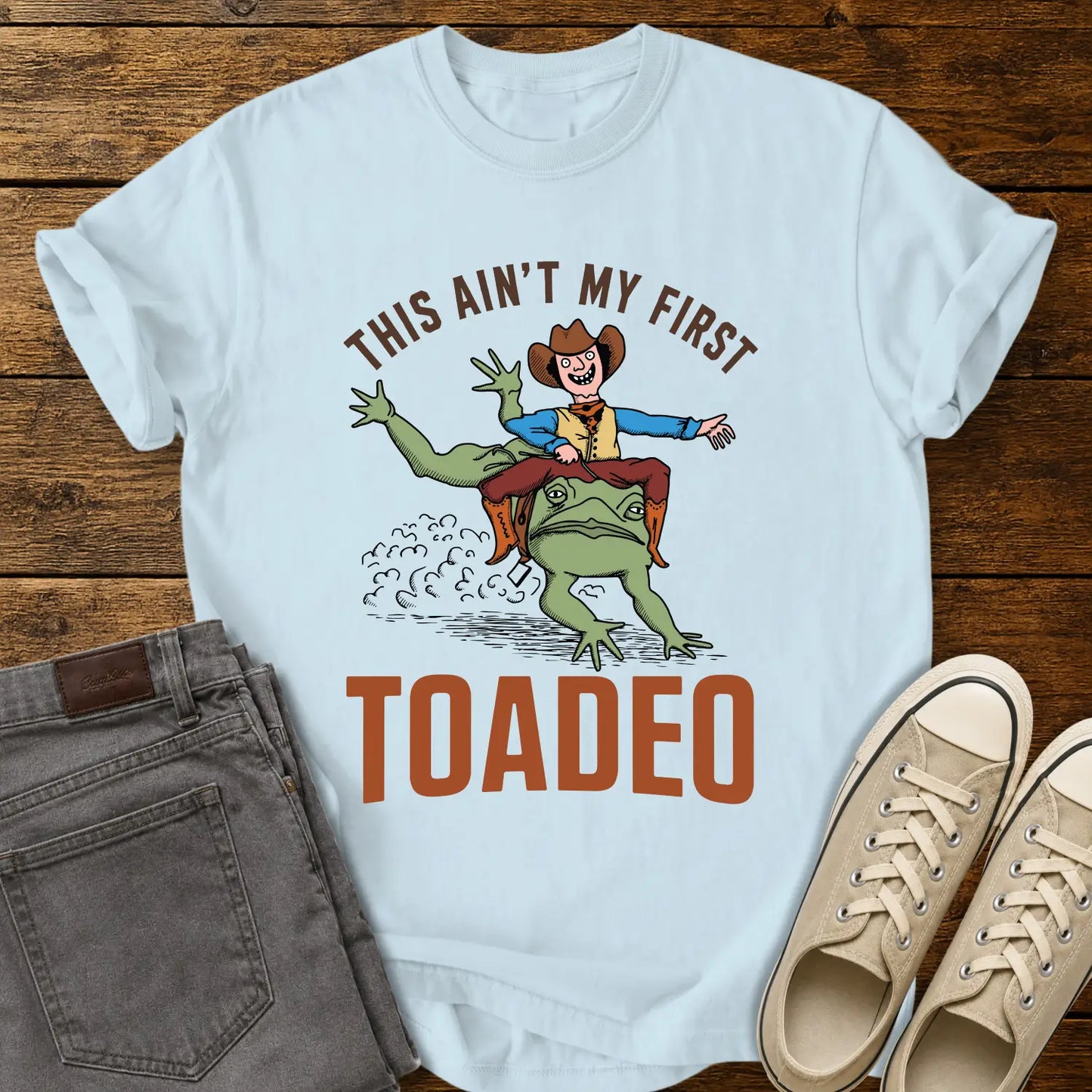Chambray blue T-shirt mockup featuring a humorous western design that reads “THIS AIN’T MY FIRST TOADEO.” The artwork shows a smiling cowboy riding a giant frog in a rodeo scene, with dust clouds behind them. The shirt is displayed on a rustic wood plank background alongside gray jeans and cream canvas sneakers.