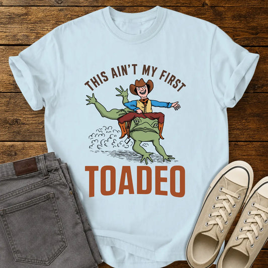 Chambray blue T-shirt mockup featuring a humorous western design that reads “THIS AIN’T MY FIRST TOADEO.” The artwork shows a smiling cowboy riding a giant frog in a rodeo scene, with dust clouds behind them. The shirt is displayed on a rustic wood plank background alongside gray jeans and cream canvas sneakers.