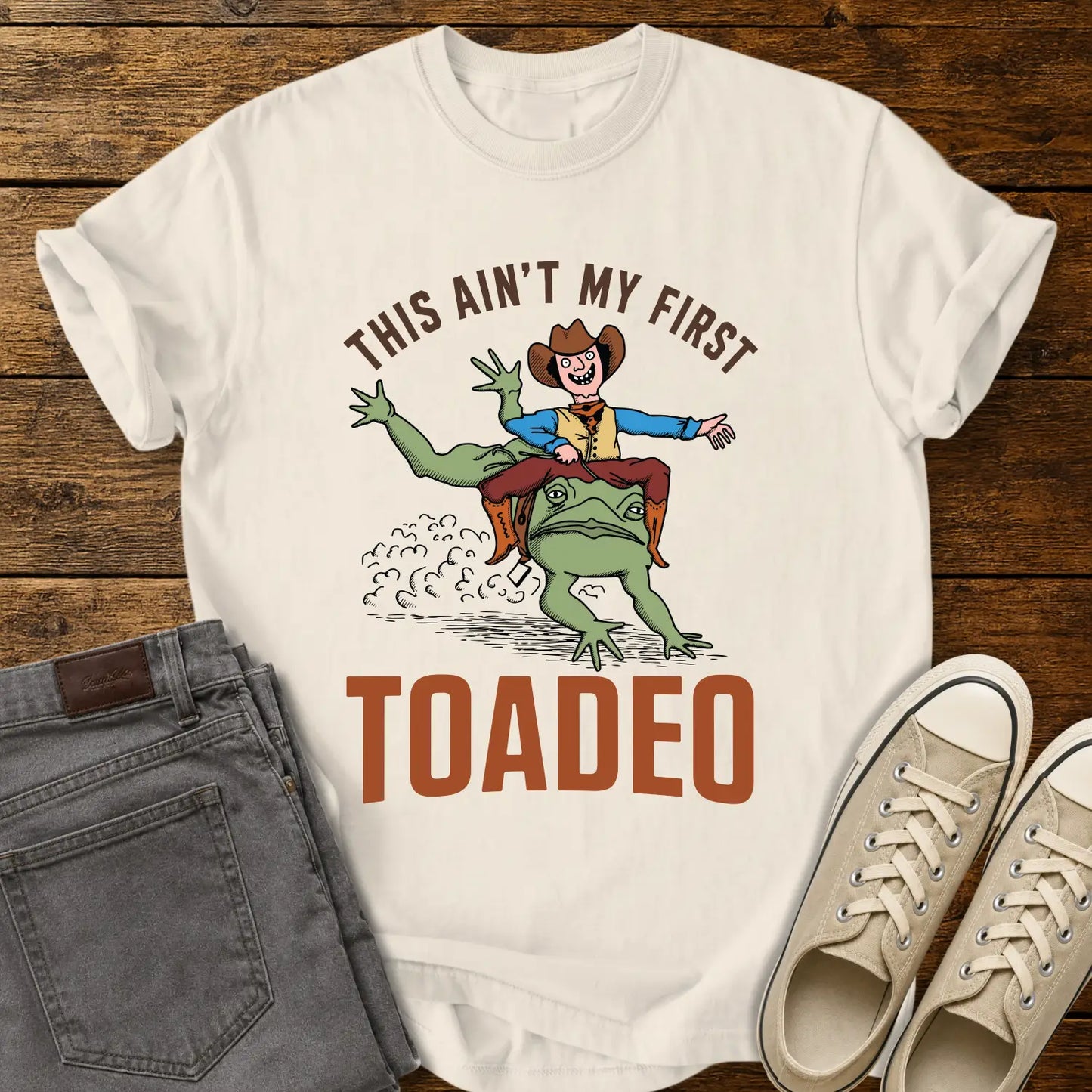 ivory T-shirt mockup featuring a humorous western design that reads “THIS AIN’T MY FIRST TOADEO.” The artwork shows a smiling cowboy riding a giant frog in a rodeo scene, with dust clouds behind them. The shirt is displayed on a rustic wood plank background alongside gray jeans and cream canvas sneakers.