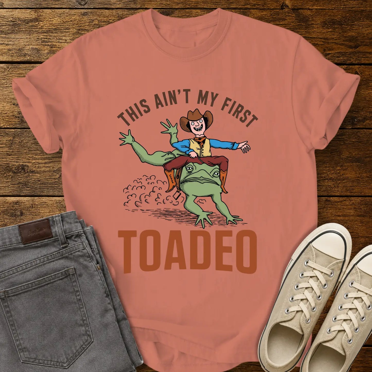 terracotta T-shirt mockup featuring a humorous western design that reads “THIS AIN’T MY FIRST TOADEO.” The artwork shows a smiling cowboy riding a giant frog in a rodeo scene, with dust clouds behind them. The shirt is displayed on a rustic wood plank background alongside gray jeans and cream canvas sneakers.