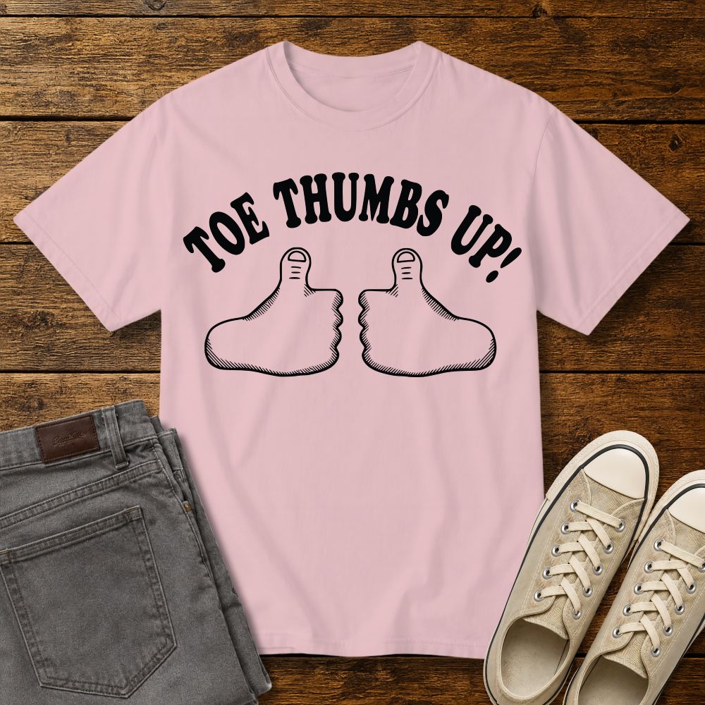 blossom pink graphic T-shirt mockup featuring two cartoon toe thumbs illustrations with the phrase “Toe Thumbs Up!” in bold black retro-style lettering, styled with gray jeans and beige sneakers on a rustic wood background.