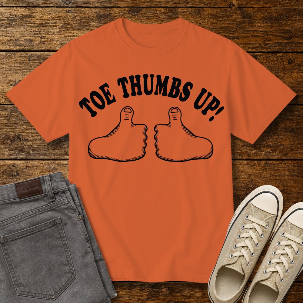 burnt orange graphic T-shirt mockup featuring two cartoon toe thumbs illustrations with the phrase “Toe Thumbs Up!” in bold black retro-style lettering, styled with gray jeans and beige sneakers on a rustic wood background.