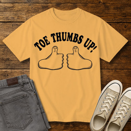 Citrus yellow graphic T-shirt mockup featuring two cartoon toe thumbs illustrations with the phrase “Toe Thumbs Up!” in bold black retro-style lettering, styled with gray jeans and beige sneakers on a rustic wood background.