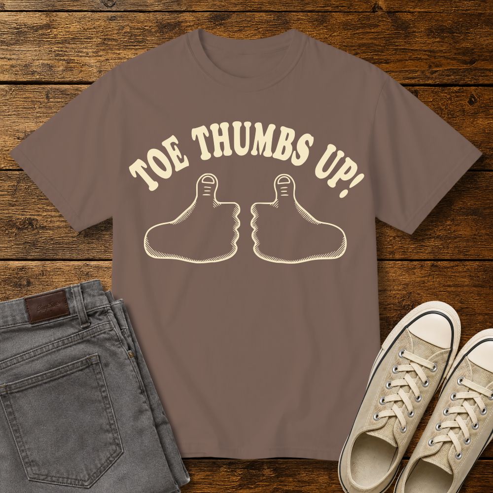 espresso brown graphic T-shirt mockup featuring two cartoon toe thumbs illustrations with the phrase “Toe Thumbs Up!” in cream retro-style lettering, styled with gray jeans and beige sneakers on a rustic wood background.