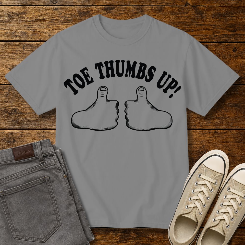 grey graphic T-shirt mockup featuring two cartoon toe thumbs illustrations with the phrase “Toe Thumbs Up!” in bold black retro-style lettering, styled with gray jeans and beige sneakers on a rustic wood background.