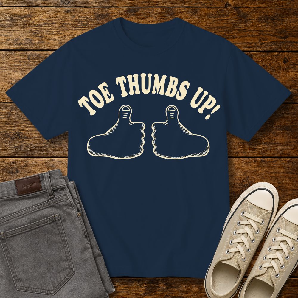 true navy graphic T-shirt mockup featuring two cartoon toe thumbs illustrations with the phrase “Toe Thumbs Up!” in cream retro-style lettering, styled with gray jeans and beige sneakers on a rustic wood background.